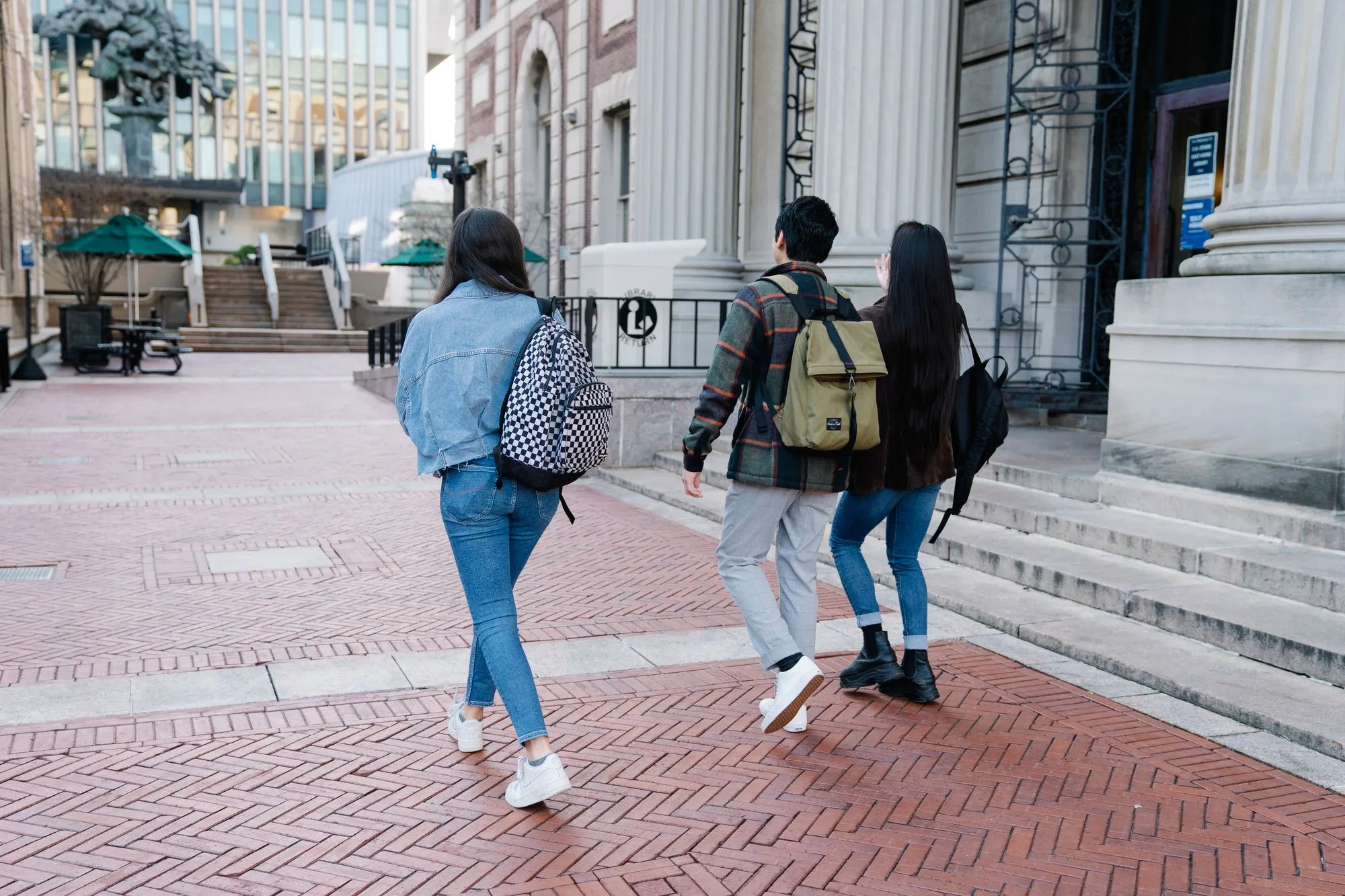 Three students with backpacks walking away from the camera across a brick campus plaza toward a university library.